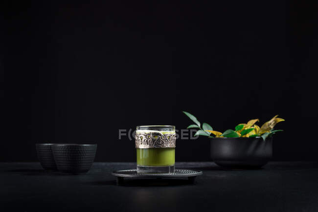 Composición de naturaleza muerta con té matcha oriental tradicional servido en taza de vidrio con decoración ornamental de metal en la mesa con cuencos de cerámica y hojas verdes frescas sobre fondo negro - foto de stock