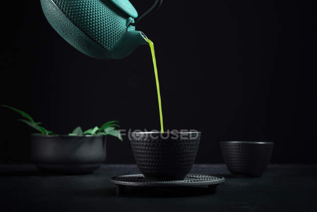 Chá matcha japonês saudável sendo derramado de bule verde em tigela de cerâmica preta durante a cerimônia de chá contra fundo preto — Fotografia de Stock