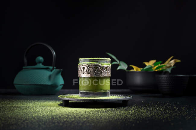 Still life composition with traditional oriental matcha tea served in glass cup with metal ornamental decor on table with ceramic bowls and fresh green leaves against black background — Stock Photo