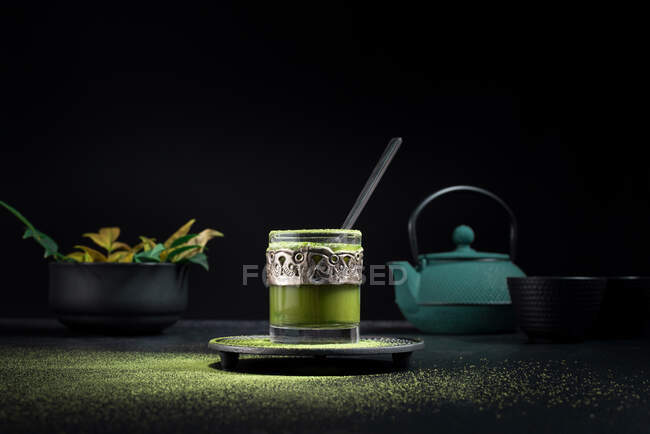 Composition nature morte avec thé matcha oriental traditionnel servi dans une tasse en verre avec décor ornemental en métal sur la table avec bols en céramique et feuilles vertes fraîches sur fond noir — Photo de stock