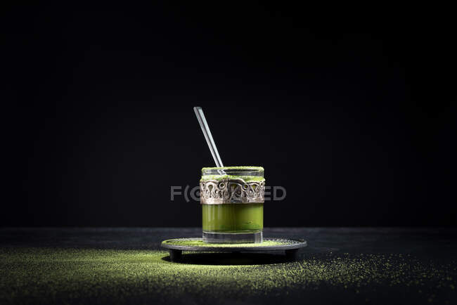 Sano tè verde matcha alle erbe servito in tazza di vetro con decorazione in metallo sul piattino cosparso di polvere sul tavolo nero — Foto stock