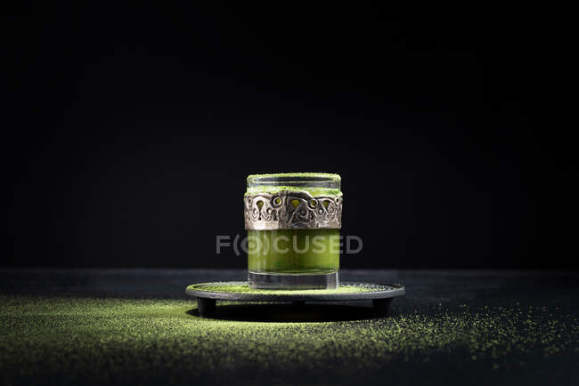 Healthy herbal green matcha tea served in glass cup with metal decoration on saucer sprinkled with powder on black table — Stock Photo