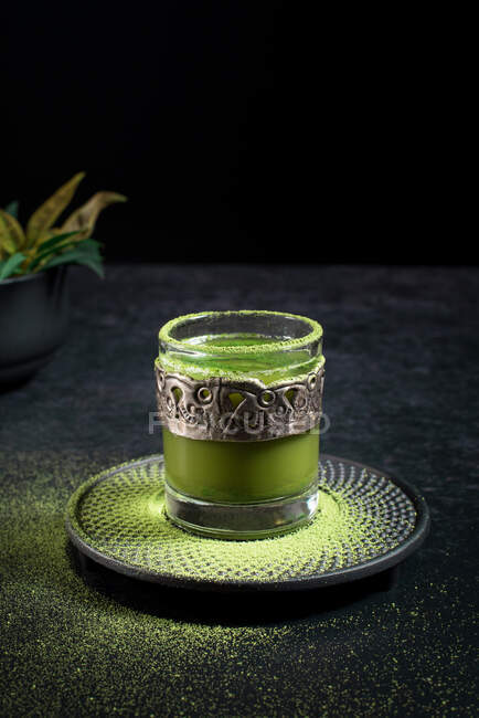 From above of healthy herbal green matcha tea served in glass cup with metal decoration on saucer sprinkled with powder on black table — Stock Photo