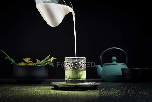 Leche fresca que se vierte del frasco en una taza de vidrio con té matcha colocado en la mesa con tetera y vajilla durante la ceremonia tradicional - foto de stock