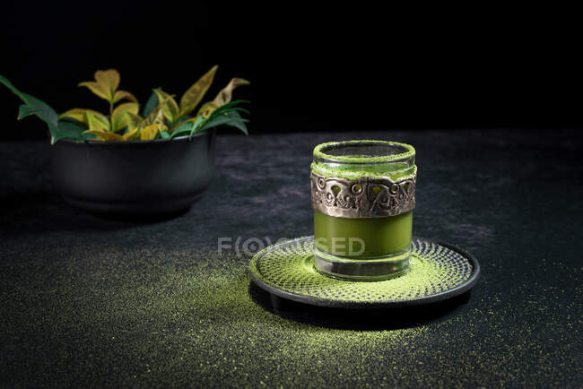 De cima de ervas saudáveis chá matcha verde servido em copo de vidro com decoração de metal no pires polvilhado com pó na mesa preta — Fotografia de Stock