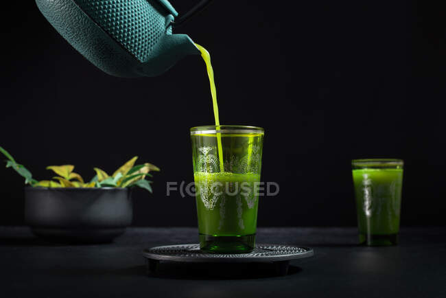 Chá matcha japonês saudável sendo derramado de bule verde em vidro durante a cerimônia do chá contra fundo preto — Fotografia de Stock