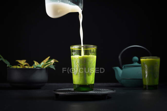 Latte fresco versato dal barattolo nella tazza di vetro con tè matcha posto sulla tavola con teiera e stoviglie durante la cerimonia tradizionale — Foto stock