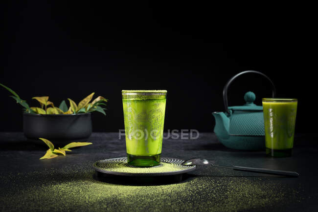 Натюрморт с традиционным восточным чаем маття подается в стеклянной чашке с металлическим декоративным декором на столе с керамическими чашами и свежими зелеными листьями на черном фоне — стоковое фото