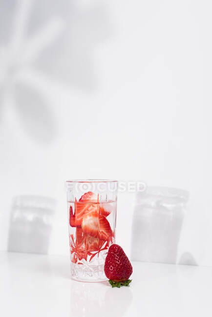 Eau saine et rafraîchissante infusée de désintoxication aux fraises fraîches coupées en tranches, servie en verre transparent contre un mur blanc ombragé — Photo de stock