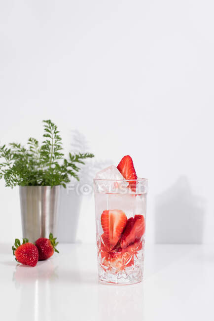 Refrescante bebida de verano con fresas frescas en rodajas y cubitos de hielo con agua servida en vidrio sobre una mesa blanca - foto de stock