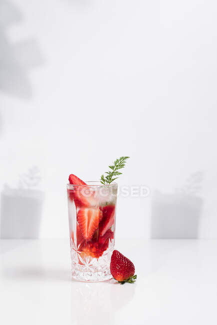 Agua sana y refrescante para desintoxicar con fresas frescas en rodajas maduras servidas en vidrio transparente contra la pared blanca con sombras - foto de stock