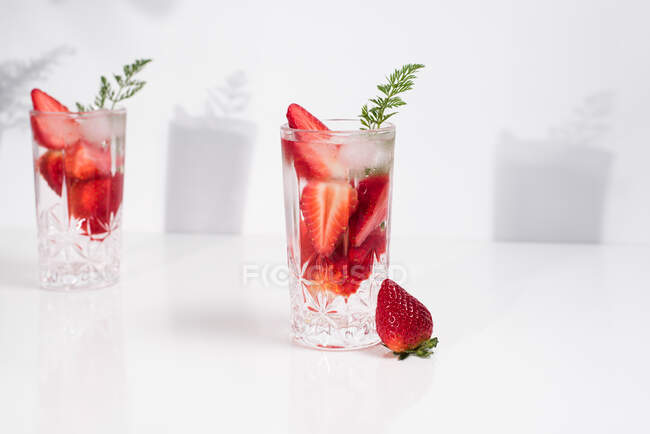 Refrescante bebida de verano con fresas frescas en rodajas y cubitos de hielo con agua servida en vasos sobre mesa blanca - foto de stock