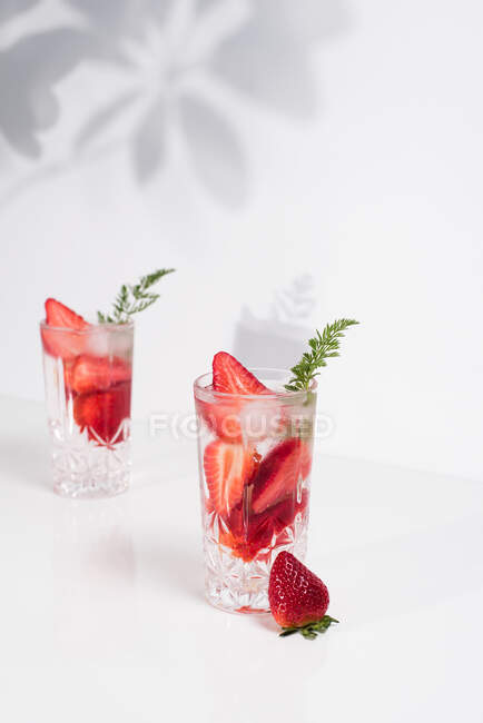 Освежающий летний напиток со свежей клубникой и кубиками льда с водой, подаваемой в стаканах на белом столе — стоковое фото