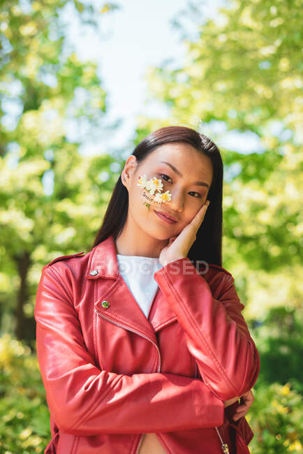Веселая этническая женщина в красной куртке с цветущими цветами на щеке, смотрящая в камеру, стоя на солнце — стоковое фото