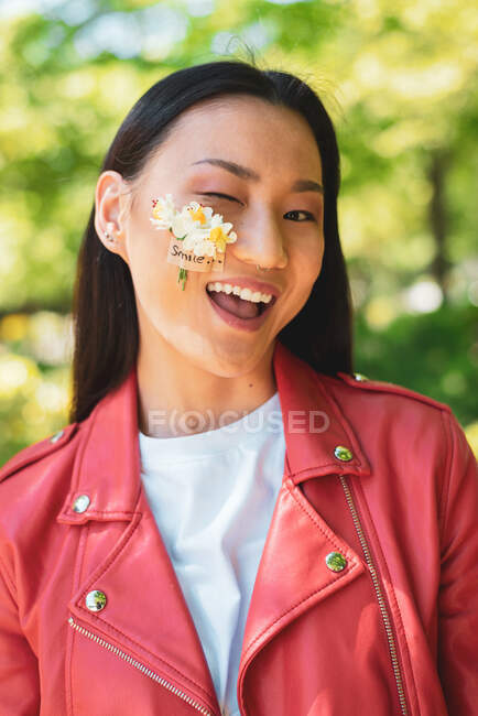 Fröhliche ethnische Frau in roter Jacke mit blühenden Blumen auf der Wange, die im Sonnenlicht zwinkernd in die Kamera blickt — Stockfoto
