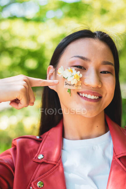 Fröhliche ethnische Frau in roter Jacke mit blühenden Blumen auf der Wange, die in die Kamera schaut, während sie im Sonnenlicht steht — Stockfoto