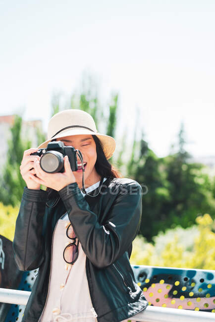 Content ethnic female in hat and leather jacket taking photo on professional camera on sunny day — Stock Photo