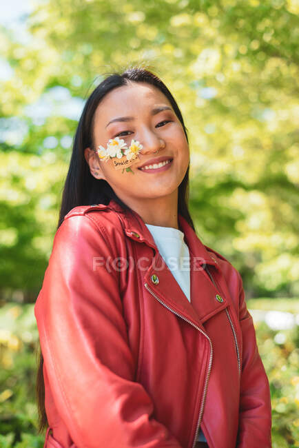 Весела етнічна жінка в червоній куртці з квітами на щоці, дивлячись на камеру, стоячи на сонячному світлі — стокове фото