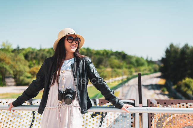 Mujer étnica sonriente en ropa elegante con cámara fotográfica digital apoyada en la cerca del puente mientras mira hacia otro lado a la luz del sol - foto de stock