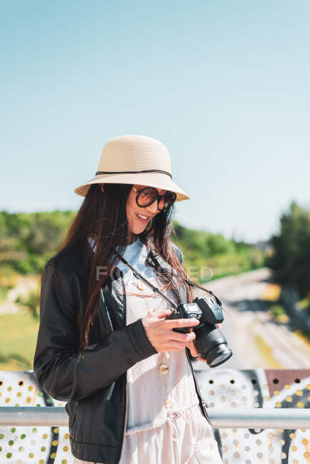 Content ethnic female in hat and leather jacket with digital photo camera standing on bridge in sunlight — Stock Photo