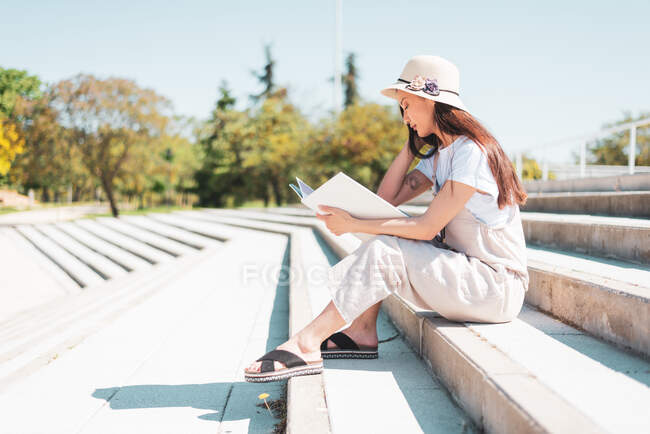Вид сбоку на молодую этническую женщину в шляпе и сандалиях, читающую учебник, сидя на лестнице на открытом воздухе — стоковое фото