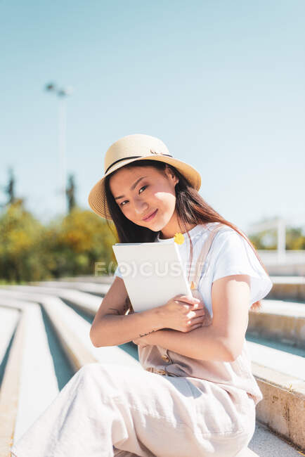 Conteúdo Asiática fêmea de chapéu com livro olhando para a câmera enquanto descansa em escadas urbanas à luz do sol — Fotografia de Stock
