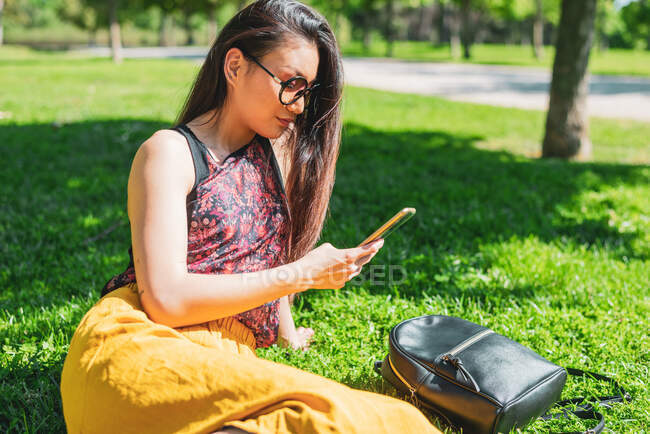 Jeune femme ethnique en lunettes messagerie texte sur téléphone portable assis sur la prairie avec sac à dos en cuir à la lumière du soleil — Photo de stock