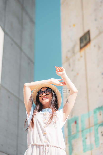 From below of content ethnic female with tattoos in trendy apparel and hat looking away in city on sunny day — Stock Photo