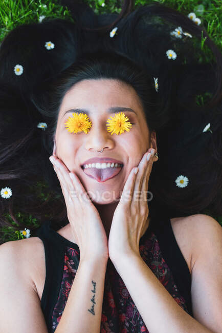 Top view of content female with tattoo and blooming flowers on eyes touching face while lying with tongue out on meadow — Stock Photo