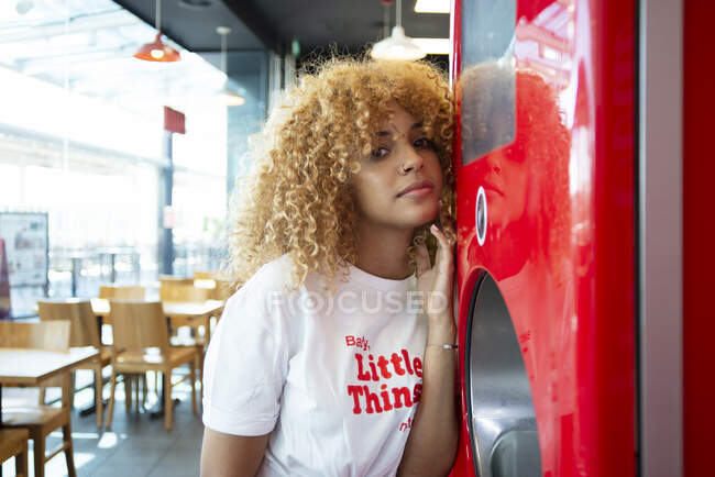 Молодая афроамериканка с вьющимися волосами, опирающаяся на красный автомат с газировкой в кафе и смотрящая в камеру — стоковое фото
