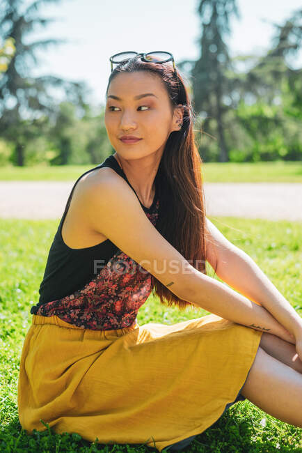 Jovem fêmea étnica macia com óculos olhando para longe sobre o ombro enquanto sentado no prado no dia ensolarado — Fotografia de Stock