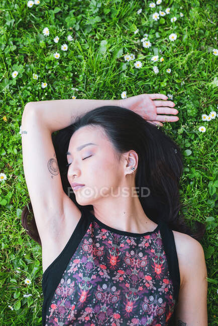 Vista aérea de hembra étnica suave en ropa ornamental acostada con los ojos cerrados en el césped con manzanillas florecientes en verano - foto de stock
