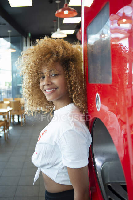Young African American female with curly hair leaning on red soda machine in cafe and looking at camera — Stock Photo