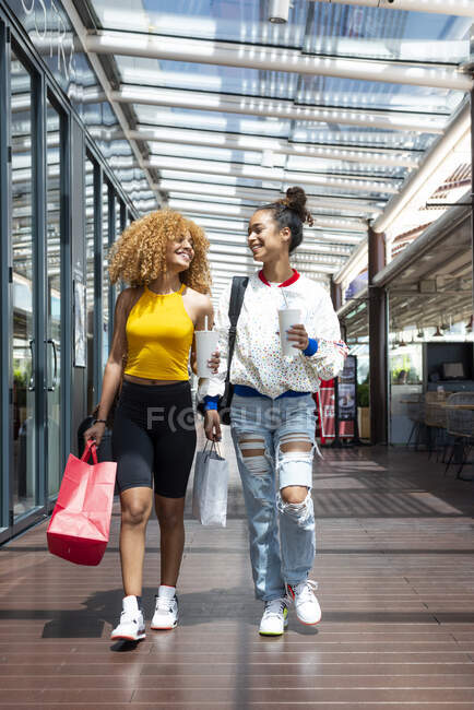 Positive African American female friends with shopping paper bags walking along mall and looking at each other — Stock Photo