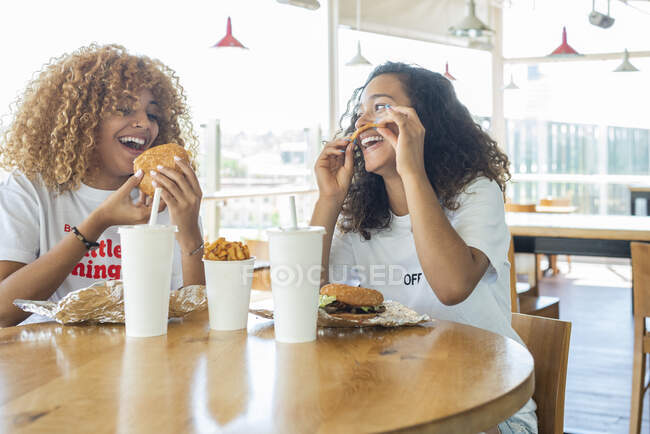 Amigas afro-americanas positivas sentadas à mesa com fast food e bebidas e desfrutando de fim de semana no café — Fotografia de Stock
