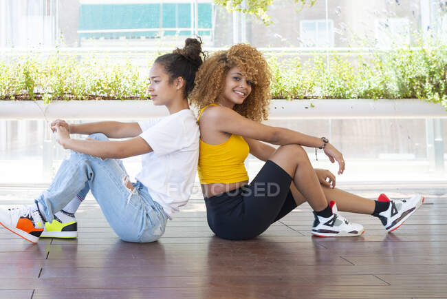 Вид сбоку на счастливых афроамериканских подруг, сидящих спиной к спине на деревянном полу на веранде — стоковое фото