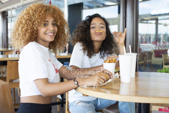 Amigos afroamericanos positivos sentados a la mesa con comida rápida y bebidas y disfrutando del fin de semana en la cafetería - foto de stock