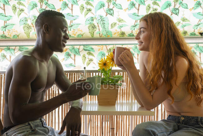 Vista lateral do casal multirracial sentado à mesa e bebendo bebidas quentes em casa durante o café da manhã — Fotografia de Stock