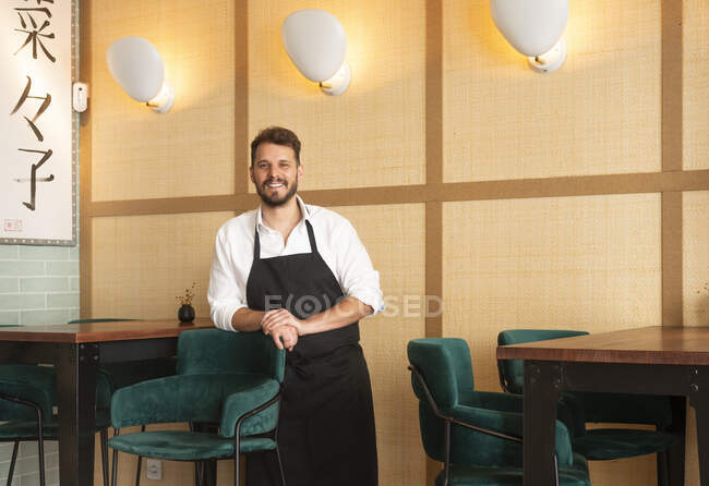 Chef masculino alegre en delantal de pie en el restaurante de sushi y mirando a la cámara - foto de stock