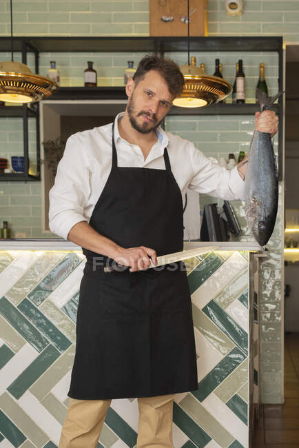 Мужчина повар в фартуке стоит с огромной рыбой и ножом в суши-ресторане и смотрит в камеру — стоковое фото