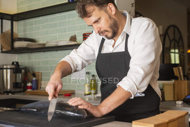 Chef masculino concentrado en delantal cortando pescado fresco crudo en la mesa en el restaurante de sushi - foto de stock