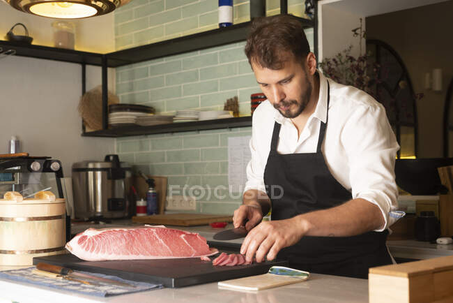 Сфокусований чоловік - кухар ріже сиру рибу за столом в азіатському ресторані і готує суші. — стокове фото