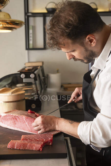 Seitenansicht eines männlichen Chefkochs, der rohen Fisch am Tisch eines asiatischen Restaurants schneidet und Sushi zubereitet — Stockfoto