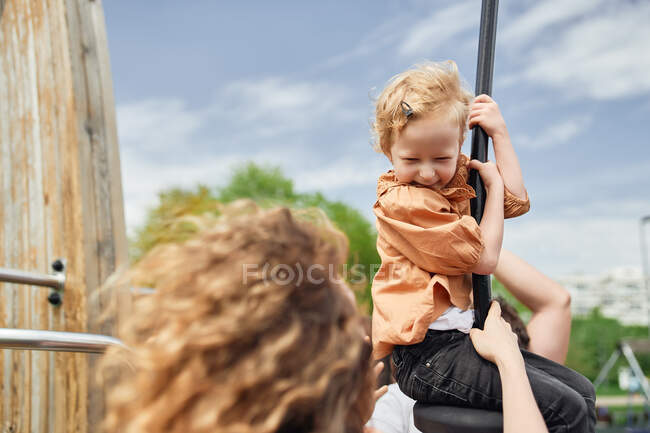 Feliz niña adorable montar columpio cuerda mientras se divierten en el patio de recreo bajo la supervisión de la madre - foto de stock