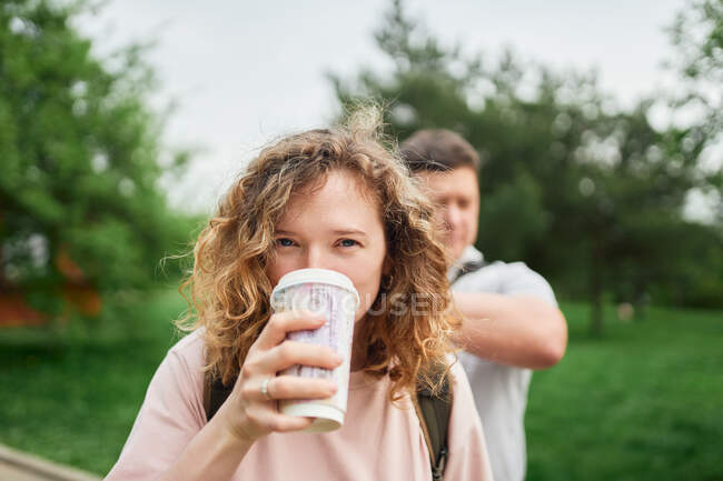 Очаровательная женщина с вьющимися волосами наслаждается горячим напитком в бумажной чашке, чтобы пойти, глядя на камеру в летнем парке — стоковое фото