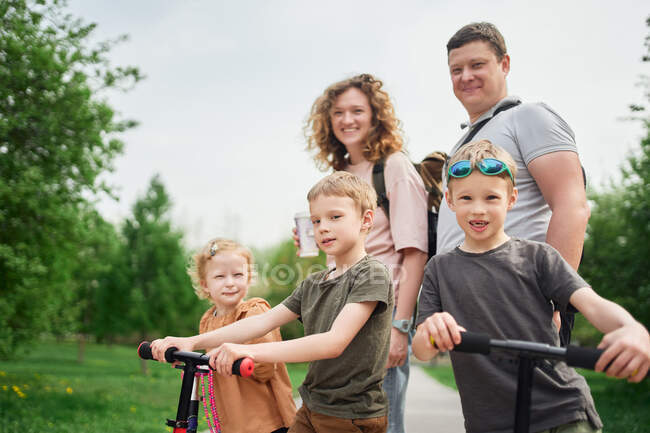 Encantada pareja con niños en scooters pasando el fin de semana juntos en el parque y mirando a la cámara - foto de stock