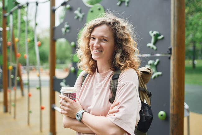 Delighted female with curly hair holding takeaway while standing on playground in park and looking at camera — Stock Photo