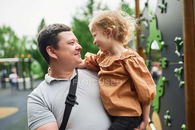 Улыбающийся отец обнимает милую дочь на детской площадке летом, глядя друг на друга — стоковое фото