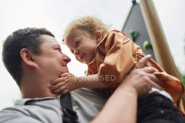 Von unten lächelnder Vater, der seine süße Tochter im Sommer auf dem Spielplatz umarmt, während er einander ansieht — Stockfoto