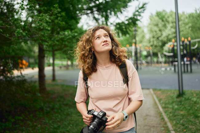 Fotógrafa enfocada con cámara moderna de pie en el parque verde y mirando hacia arriba - foto de stock